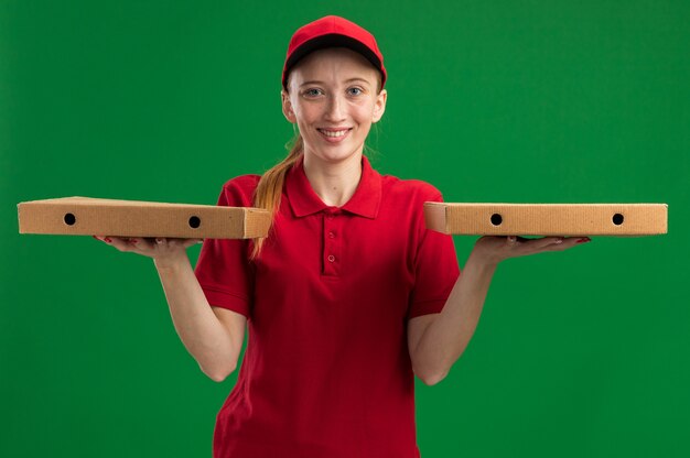 Repartidor joven en uniforme rojo y gorra sosteniendo cajas de pizza con una sonrisa en la cara feliz de pie sobre la pared verde