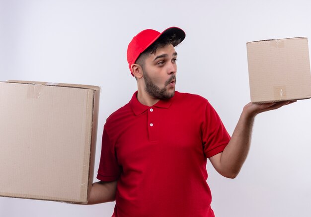 Repartidor joven en uniforme rojo y gorra sosteniendo grandes cajas de cartón que parecen desconcertados tratando de tomar una decisión