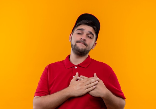 Repartidor joven en uniforme rojo y gorra sosteniendo las manos cruzadas sobre el pecho con los ojos cerrados sintiendo emociones positivas