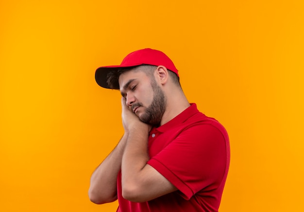 Repartidor joven con uniforme rojo y gorra sosteniendo las palmas juntas con la cabeza apoyada en las palmas quiere dormir cansado y con exceso de trabajo