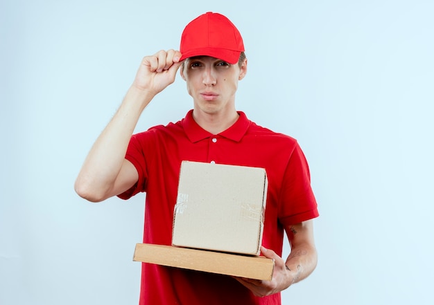 Repartidor joven en uniforme rojo y gorra sosteniendo el paquete de caja y caja de pizza mirando confiado tocando su gorra de pie sobre la pared blanca