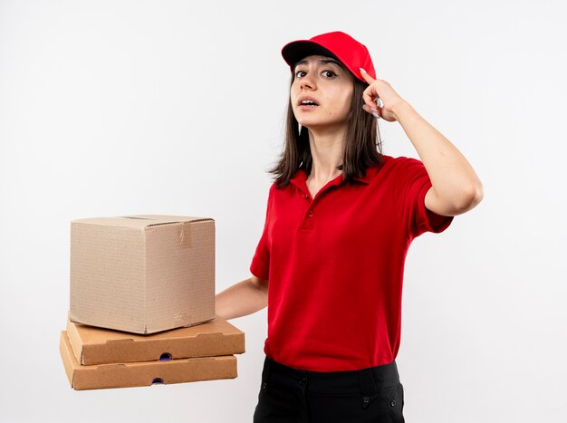 Repartidor joven con uniforme rojo y gorra sosteniendo el paquete de caja y cajas de pizza apuntando con el dedo índice a su sien luciendo confiada concentrada en una tarea de pie sobre una pared blanca