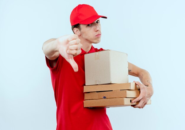 Repartidor joven en uniforme rojo y gorra sosteniendo el paquete de caja y cajas de pizza mirando al frente disgustado mostrando tumbs de pie sobre una pared blanca