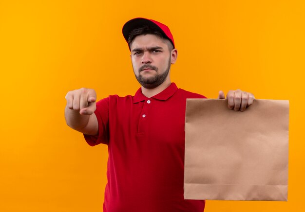 Repartidor joven en uniforme rojo y gorra sosteniendo el paquete de papel apuntando con el dedo a la cámara con cara enojada
