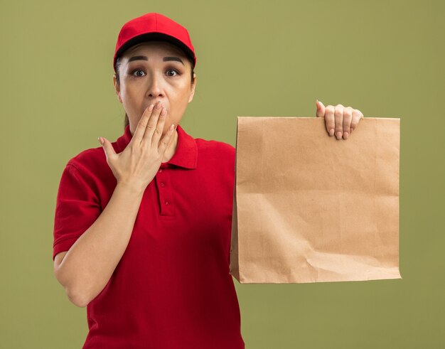 Repartidor joven en uniforme rojo y gorra sosteniendo el paquete de papel conmocionado cubriendo la boca con la mano