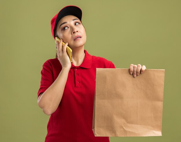 Repartidor joven en uniforme rojo y gorra sosteniendo el paquete de papel mirando confundido mientras habla por teléfono móvil de pie sobre la pared verde