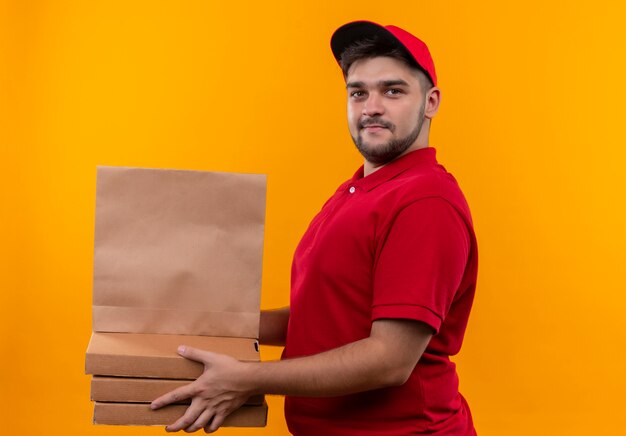 Repartidor joven en uniforme rojo y gorra sosteniendo el paquete de papel y la pila de cajas de pizza que parecen seguros