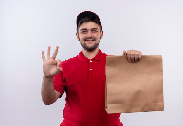 Repartidor joven en uniforme rojo y gorra sosteniendo el paquete de papel sonriendo amigablemente mostrando el signo de ok