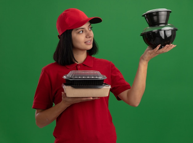 Repartidor joven en uniforme rojo y gorra sosteniendo paquetes de alimentos mirando confiado sonriendo de pie sobre la pared verde