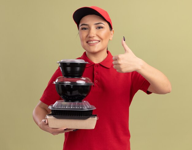 Repartidor joven en uniforme rojo y gorra sosteniendo una pila de paquetes de alimentos mirando al frente sonriendo alegremente mostrando los pulgares para arriba de pie sobre la pared verde