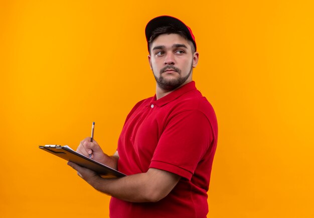 Repartidor joven en uniforme rojo y gorra sosteniendo portapapeles con lápiz mirando a un lado con cara seria