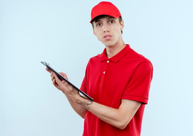 Repartidor joven en uniforme rojo y gorra sosteniendo portapapeles con páginas en blanco mirando confiado parado sobre la pared blanca