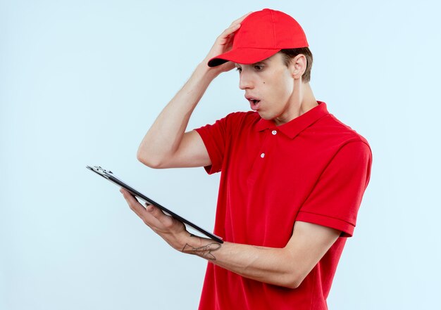 Repartidor joven en uniforme rojo y gorra sosteniendo portapapeles con páginas en blanco mirando confundido y sorprendido de pie sobre la pared blanca