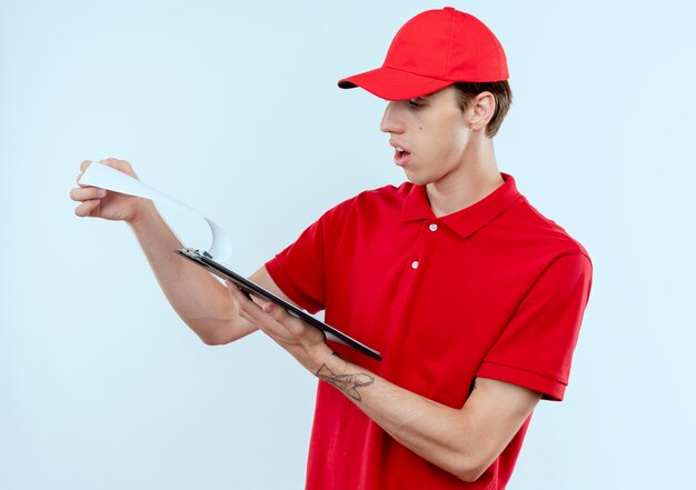 Repartidor joven en uniforme rojo y gorra sosteniendo portapapeles con páginas en blanco mirando sorprendido de pie sobre la pared blanca