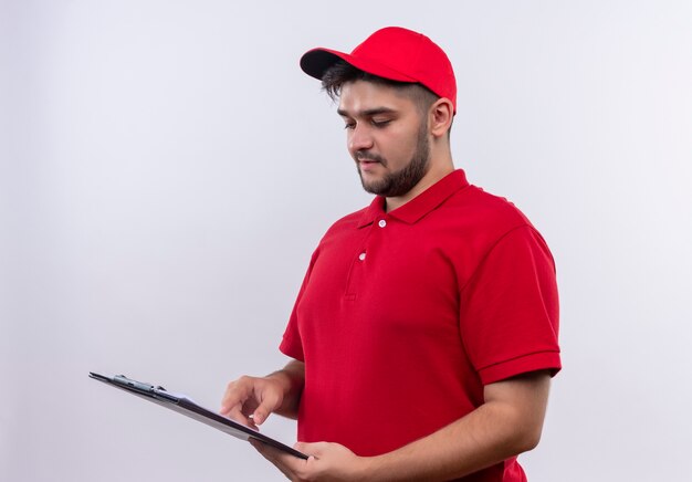 Repartidor joven en uniforme rojo y gorra sosteniendo portapapeles con páginas en blanco mirándolos con cara seria