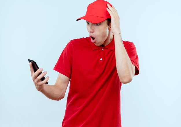 Repartidor joven con uniforme rojo y gorra sosteniendo teléfono inteligente mirando sorprendido y confundido con la mano en la cabeza por error de pie sobre la pared blanca