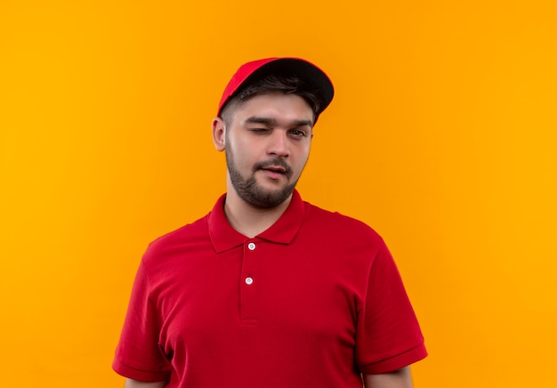 Foto gratuita repartidor joven en uniforme rojo y gorra wiking y sonriendo mirando a la cámara