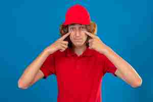 Foto gratuita repartidor joven en uniforme rojo haciendo un gesto de ser cuidadoso con las manos apuntando a sus ojos sobre fondo azul aislado