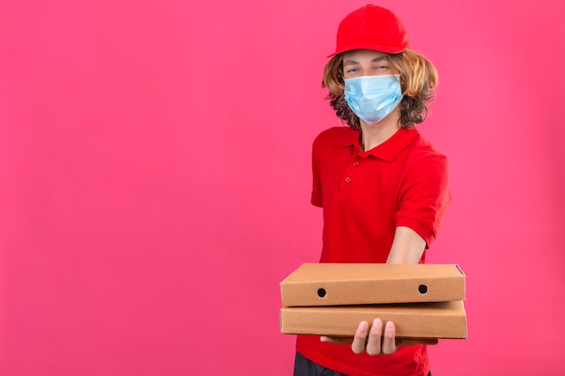 Repartidor joven en uniforme rojo con máscara médica estirando cajas de pizza sonriendo amable parado sobre fondo rosa aislado con espacio de copia