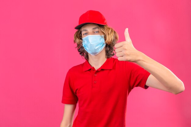 Repartidor joven en uniforme rojo con máscara médica mirando a la cámara sonriendo alegremente mostrando el pulgar hacia arriba sobre fondo rosa aislado