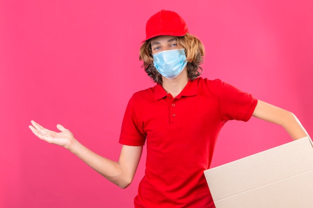 Repartidor joven en uniforme rojo con máscara médica sosteniendo una caja de cartón sonriendo alegre presentando y apuntando con la palma de la mano mirando a la cámara sobre fondo rosa aislado