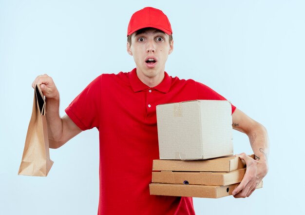 Repartidor joven en uniforme rojo y paquetes de caja de gorra mirando al frente confundido parado sobre la pared blanca