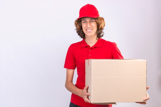 Repartidor joven en uniforme rojo sosteniendo una caja de cartón grande mirando a la cámara sonriendo alegremente de pie sobre fondo blanco aislado