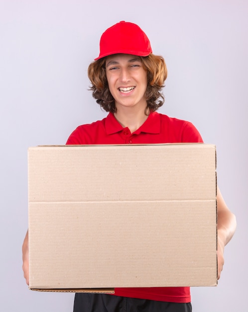 Repartidor joven en uniforme rojo sosteniendo una caja de cartón grande mirando a la cámara sonriendo alegremente de pie sobre fondo blanco aislado