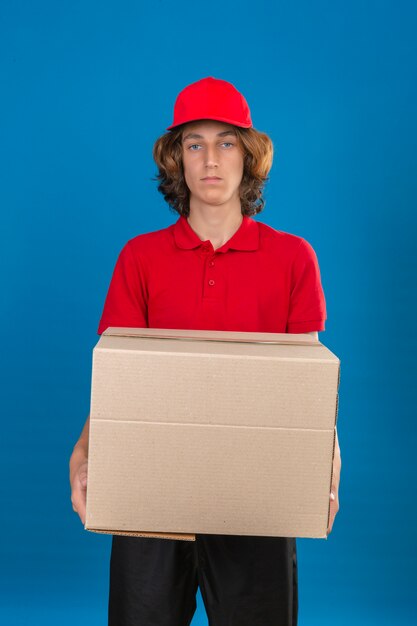 Repartidor joven en uniforme rojo sosteniendo con caja de cartón mirando a la cámara con cara seria sobre fondo azul aislado