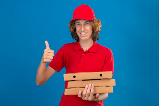 Repartidor joven en uniforme rojo sosteniendo cajas de pizza sonriendo con cara feliz mostrando el pulgar hacia arriba sobre fondo azul aislado