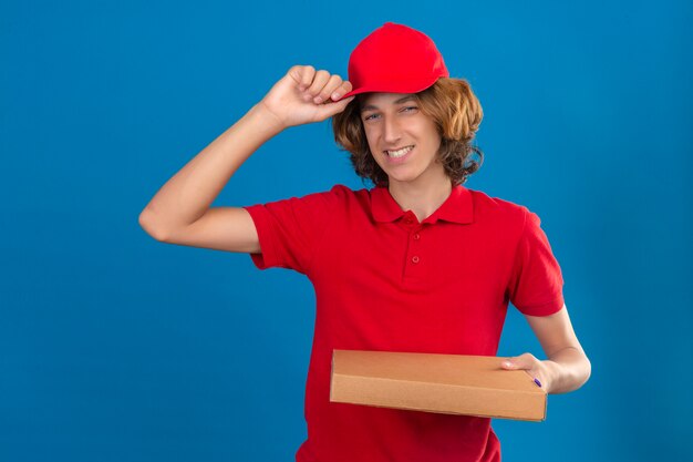 Repartidor joven en uniforme rojo sosteniendo cajas de pizza tocando su gorra en saludo sonriendo amable sobre fondo azul aislado