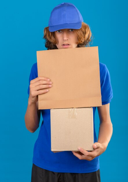 Repartidor joven vestido con camisa polo azul y gorra con caja de cartón y paquete de papel mirando sorprendido de pie sobre fondo azul aislado