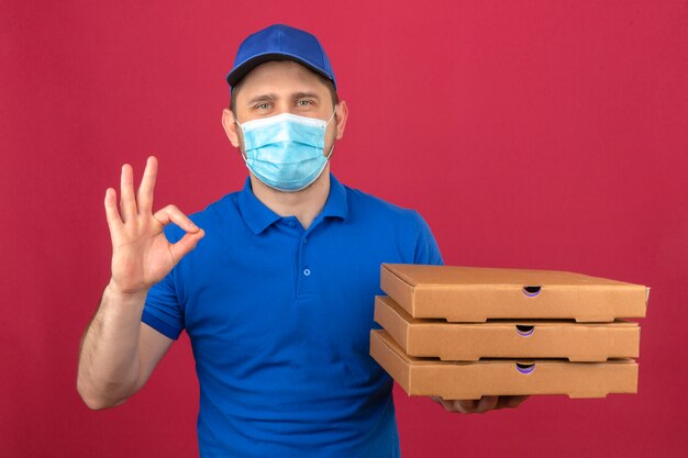 Repartidor joven vestido con camisa polo azul y gorra en máscara médica sosteniendo una pila de cajas de pizza mostrando signo de ok con cara feliz sobre fondo rosa aislado