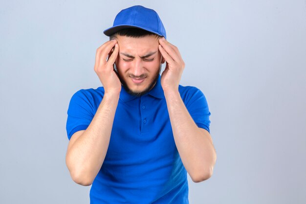 Repartidor joven vestido con camisa polo azul y gorra mirando mal tocar la cabeza que sufre de un fuerte dolor de cabeza sobre fondo blanco aislado