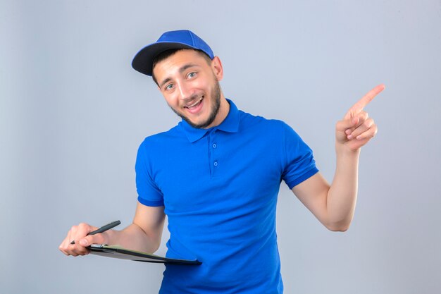 Repartidor joven vestido con camisa polo azul y gorra de pie con lápiz y apuntando hacia el lado con el dedo sobre fondo blanco aislado