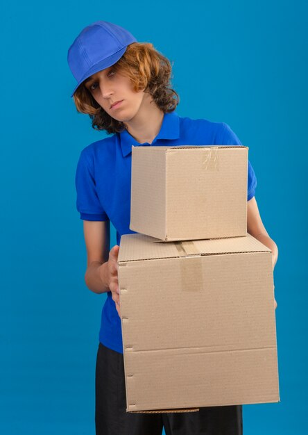 Repartidor joven vestido con camisa polo azul y gorra sosteniendo cajas de cartón mirando a la cámara con rostro serio de pie sobre fondo azul aislado