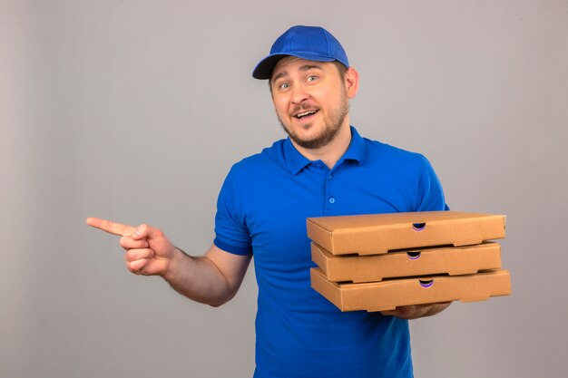 Repartidor joven vestido con camisa polo azul y gorra sosteniendo la pila de cajas de pizza apuntando hacia el lado con el dedo sonriendo alegremente sobre fondo azul aislado