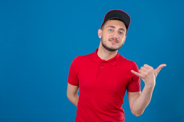 Repartidor joven vestido con camisa polo roja y gorra haciendo gesto de llamarme mirando confiado sobre fondo azul aislado