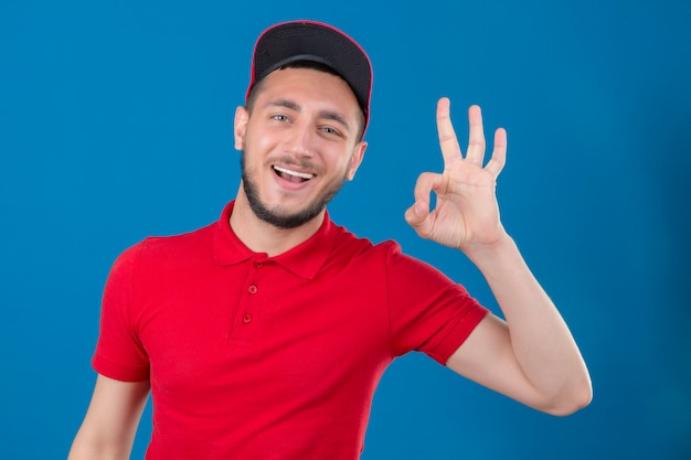 Repartidor joven vestido con camisa polo roja y gorra sonriendo alegremente haciendo bien firmar sobre fondo azul aislado