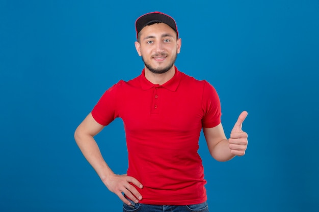 Repartidor joven vestido con polo rojo y gorra mirando a la cámara con una sonrisa amistosa mostrando el pulgar hacia arriba sobre fondo azul aislado