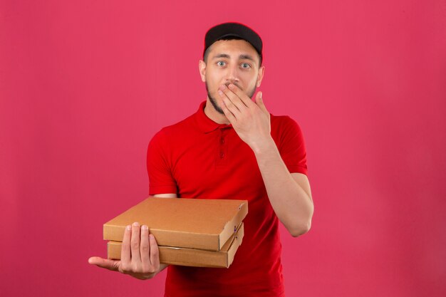 Repartidor joven vestido con polo rojo y gorra de pie con cajas de pizza mirando sorprendido y conmocionado cubriendo la boca sobre fondo rosa aislado