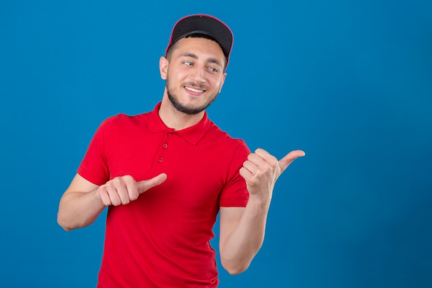 Repartidor joven vestido con polo rojo y gorra sonriendo con cara feliz mirando y apuntando hacia el lado con los pulgares hacia arriba sobre fondo azul aislado