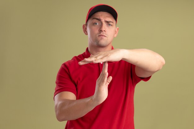 Repartidor joven vestido con uniforme y gorra mostrando gesto de tiempo de espera aislado en la pared verde oliva