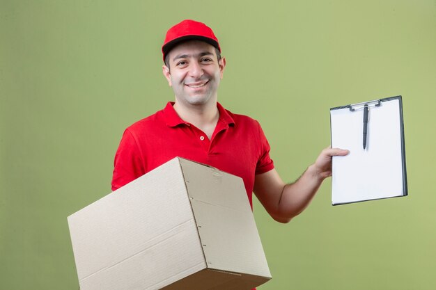 Repartidor joven vestido con uniforme rojo con caja de cartón y portapapeles mirando a la cámara con una sonrisa en la cara sobre fondo verde aislado