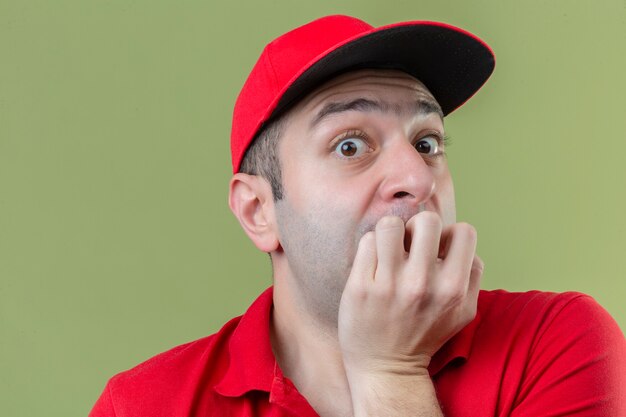 Repartidor joven vestido con uniforme rojo que parece estresado y nervioso con las manos en la boca mordiéndose las uñas sobre fondo verde aislado