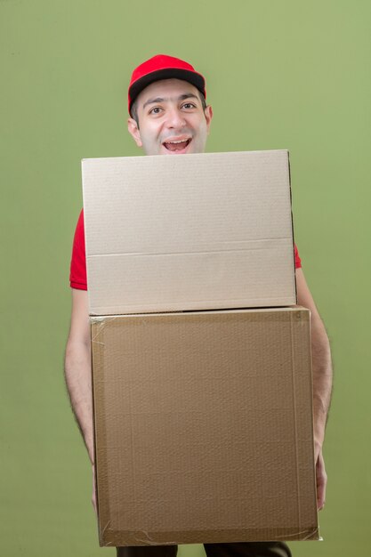 Foto gratuita repartidor joven vestido con uniforme rojo sosteniendo grandes cajas de cartón sonriendo alegremente mirando a la cámara sobre fondo verde aislado