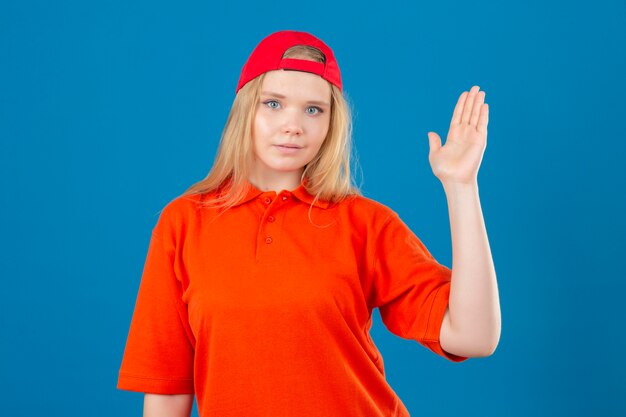 Repartidor joven vistiendo camisa polo naranja y gorra roja sonriendo amistosamente saludando con la mano dándote la bienvenida y saludándote o despidiéndote sobre fondo azul aislado