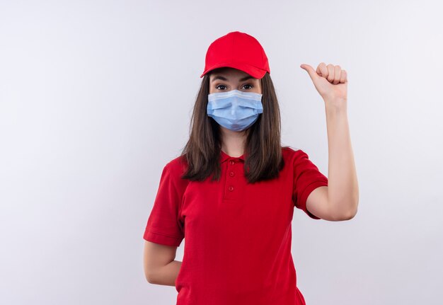 Repartidor joven vistiendo camiseta roja en gorra roja lleva mascarilla mostrando los pulgares hacia arriba sobre fondo blanco aislado