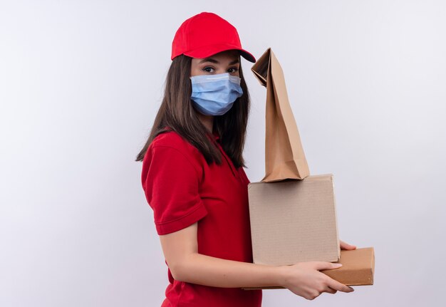 Repartidor joven vistiendo camiseta roja en gorra roja lleva mascarilla sosteniendo una caja y caja de pizza y paquete sobre fondo blanco aislado