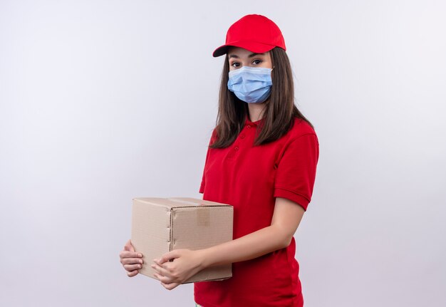 Repartidor joven vistiendo camiseta roja en gorra roja lleva mascarilla sosteniendo una caja sobre fondo blanco aislado